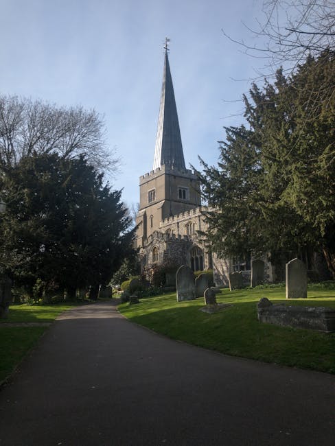 The image shows a paved pathway leading through a churchyard with several gravestones visible on both sides. Tall, mature trees with sparse branches line the pathway, partially obscuring the view of a historic stone church with a tall, pointed spire topped by a cross. The church features arched windows and stone detailing typical of traditional church architecture. In the background, a clear sky can be seen with some light cloud cover. The scene is well-lit, suggesting daytime, and the setting appears peaceful and well-maintained, suitable for a location where house removals or relocation vehicles might load or unload near a church or community area. This scene exemplifies a typical exterior environment relevant to property moves in the Bromley area, as covered by Man with Van St Mary Cray, noting the importance of accessible locations for furniture transport during home relocation.