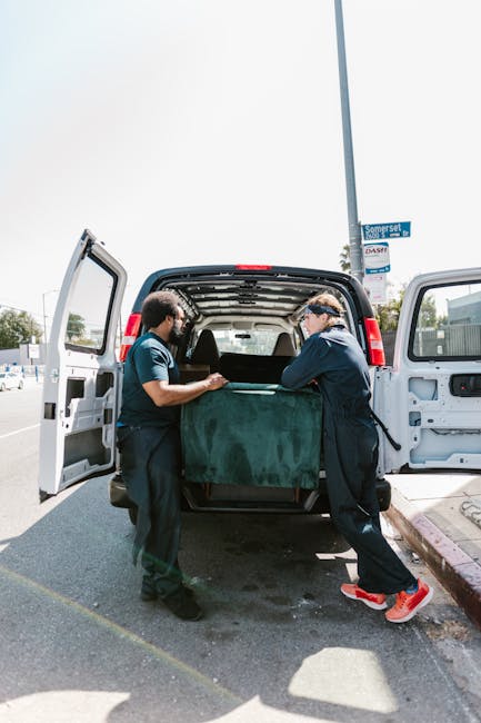 Two movers from Man with Van St Mary Cray are loading large cardboard boxes into a white delivery van parked outside a modern building with glass panels. One mover, wearing gloves and a dark jacket, i