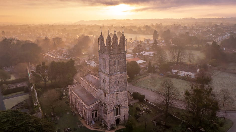 An aerial view of a historic stone church with a tall, ornate bell tower, situated in a rural landscape during sunset, with trees and fields surrounding the building. The scene shows the church's detailed Gothic architecture, with large arched windows and decorative spires at the top of the tower. In the foreground, part of the church's garden and a small graveyard with tombstones are visible. The background includes a mix of open fields, scattered trees, and distant buildings, illuminated by warm, golden light from the setting sun, creating a peaceful atmosphere that reflects the context of home or property relocation. This image exemplifies the importance of careful packing and moving logistics when managing historic or valuable properties and can be associated with professional removals and furniture transport services offered by Man with Van St Mary Cray.