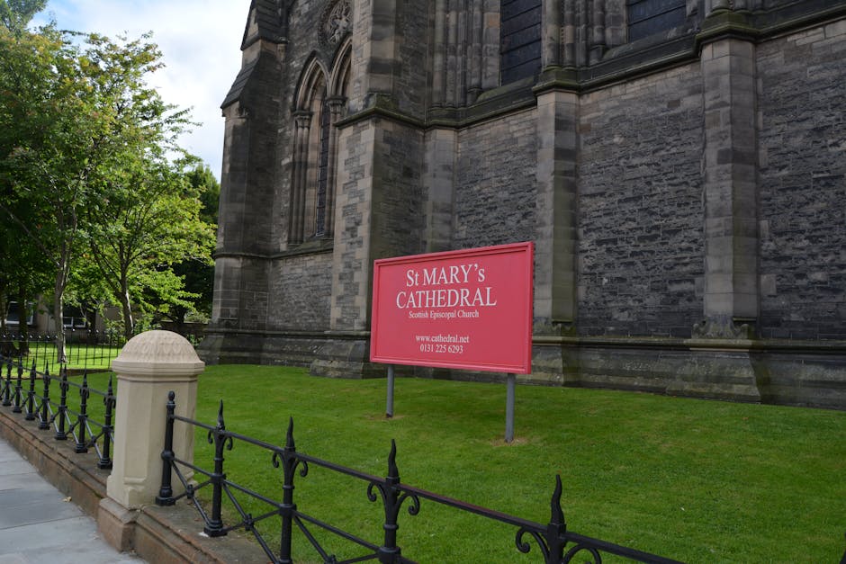 External view of St Mary’s Cathedral, a large historic church built with dark grey stone blocks featuring tall Gothic windows and detailed architectural elements. A pink signboard with white text reading 'St MARY'S CATHEDRAL, Scottish Episcopal Church' is positioned on a well-maintained grassy area next to a stone pillar and a decorative black metal fence. The sign also displays a website URL and contact number. The surrounding environment includes leafy green trees and a paved sidewalk along the church exterior, suggesting a peaceful, well-kept setting. The scene is illuminated by natural daylight, indicating clear weather, and depicts the exterior of a prominent religious building possibly undergoing or preparing for a house or furniture relocation involving logistical planning and packing, as managed by companies such as Man with Van St Mary Cray.