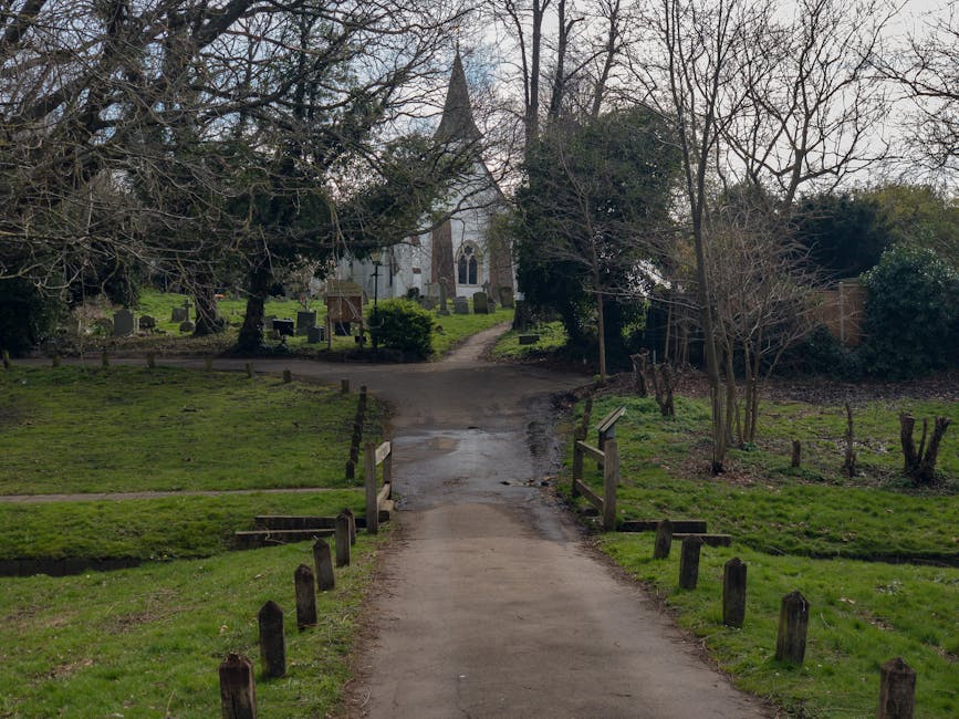 A narrow, paved pathway leading through green grass and trees towards a small church with a pointed spire, situated in Foots Cray Meadows. The path is bordered by wooden posts and low fencing on both sides, with some muddy patches visible along its surface. In the background, a historic stone church with arched windows and a steeple is partially obscured by leafless and evergreen trees, suggesting a winter or early spring season. The area around the church contains small grave markers and headstones, covered by well-maintained grass and shrubs. The scene is outdoors, illuminated by natural daylight with overcast sky conditions, capturing the peaceful, rural setting typical of a peaceful home relocation or furniture transport process near Foots Cray Meadows, as facilitated by Man with Van St Mary Cray.