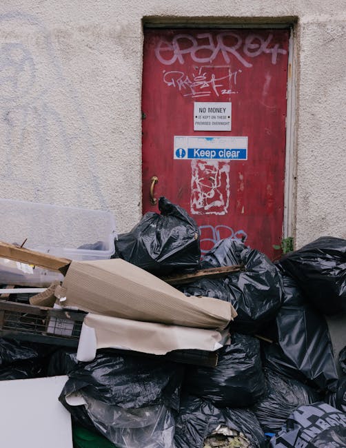 A pile of black rubbish bags, cardboard boxes, and packaging materials positioned on the pavement against a textured, off-white building wall with a red metal door in the background. The door features