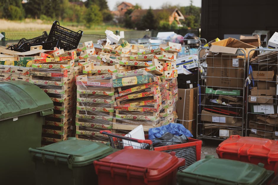 A pile of black rubbish bags, cardboard boxes, and packaging materials positioned on the pavement against a textured, off-white building wall with a red metal door in the background. The door features