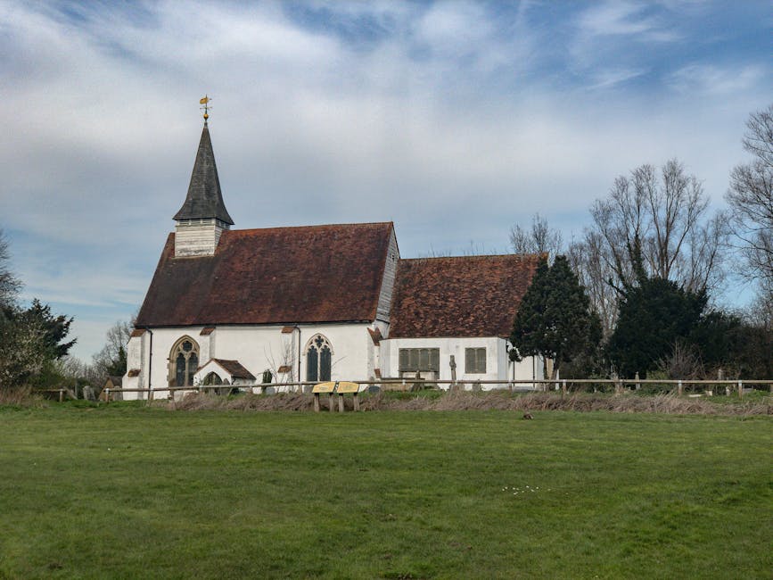 A white, traditional church with a steeply pitched, red-tiled roof and a tall, pointed steeple topped with a weather vane, situated in a rural setting. The church features arched windows with stained glass, and there are leafless trees surrounding it. In the foreground, a well-maintained grassy area extends to a wooden fence that runs along the property boundary. The sky above is partly cloudy with patches of blue. The scene provides an exterior view of the church, which could be part of a house relocation or community move context, reflecting the type of local building often found in residential areas such as St Mary Cray BR5, relevant to house removals and moving services. The image includes no furniture, boxes, vehicles, or loading equipment visible, focusing solely on the church building and nearby landscape.