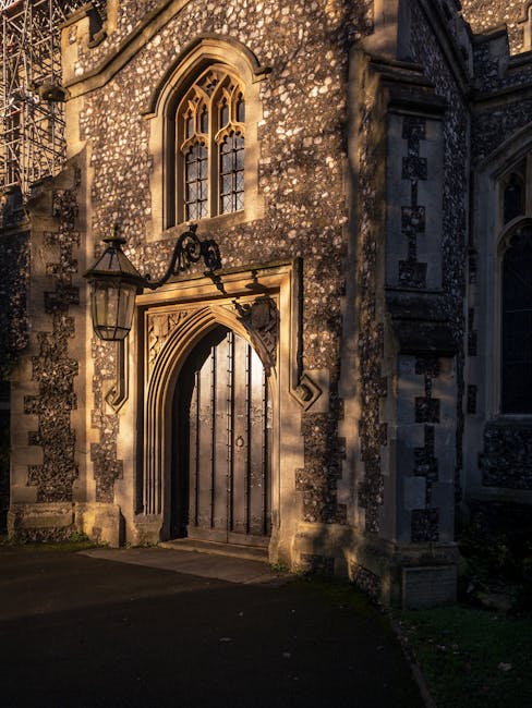 The image shows the exterior of a historic stone building with an arched wooden doorway featuring decorative carvings and a small ornamental sculpture above it. To the left of the doorway, there is an old-fashioned lantern hanging from a wrought iron bracket, casting a warm glow on the textured stone facade. Above the door, an arched window with leaded glass panes is visible, complemented by another window with similar design elements further above. The building's stonework includes dark and light patches, suggesting age and character. Shadows and sunlight create dappled patterns across the surface, highlighting the architectural details. The setting appears to be at dusk or dawn, with soft, natural lighting. The scene conveys a historic, well-preserved structure that could be part of a church, manor, or similar heritage property, ideal for a house relocation or moving services context. Man with Van St Mary Cray may occasionally operate in areas with historic buildings, assisting with furniture transport and packing during home removals.