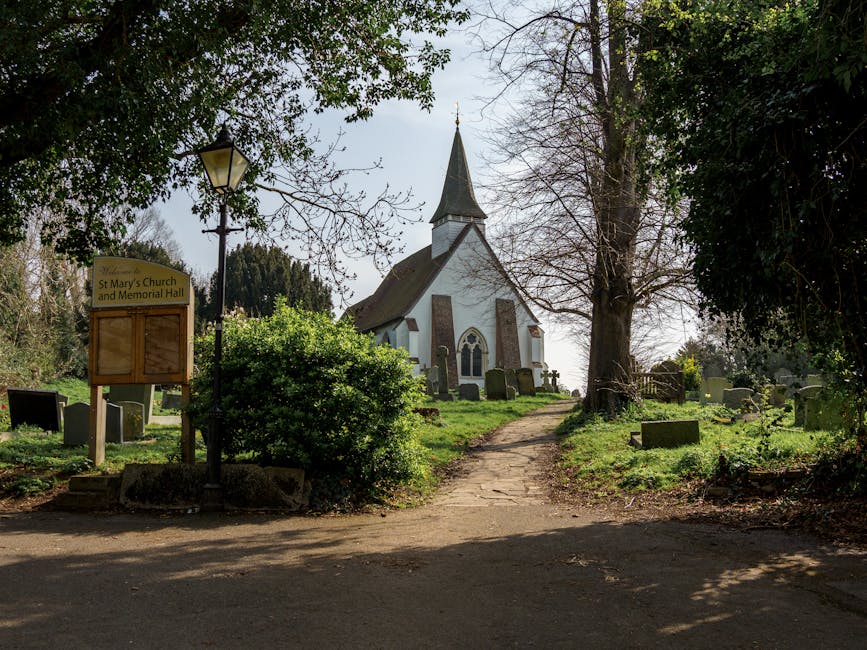 A paved pathway leading through a churchyard with grass and scattered gravestones on either side, towards a white church building with a steep, dark spire visible against a partly cloudy sky. The churchyard is surrounded by trees, some of which are leafless while others have dense green foliage. To the left of the pathway, there is a sign indicating 'St Mary's Church and Memorial Hall,' attached to a wooden post, alongside a vintage-style lamp post. The scene is outdoors during daylight, with natural light illuminating the area, which is typical for a setting involved in house removals or packing and moving activities managed by Man with Van St Mary Cray. The surrounding environment suggests a peaceful residential or community area near a historic church, with objects such as gravestones and greenery visible in the background.