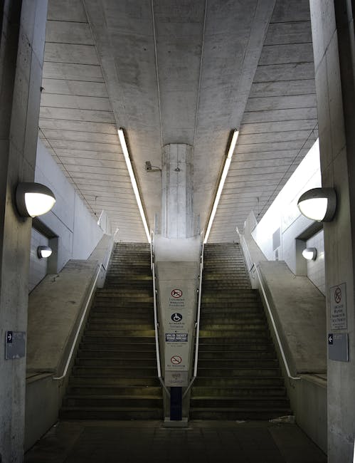 The image shows a symmetrical concrete staircase and escalator leading up from an underground station at St Mary Cray. The stairs are divided by a central column with safety signage, including no-smoking and no-alcohol symbols. On each side of the escalator, there are smooth, angled concrete walls with integrated modern wall-mounted lights providing illumination. Overhead, fluorescent strip lighting runs along the ceiling, highlighting the utilitarian design of the station's interior. The environment appears clean and well-maintained, emphasizing the functional layout of the station. This setting exemplifies a typical urban transit environment where individuals may transfer to and from station exits, supporting logistical considerations for house removals and furniture transport arranged with [COMPANY_NAME], such as loading and unloading within station areas during home relocation or moving processes.