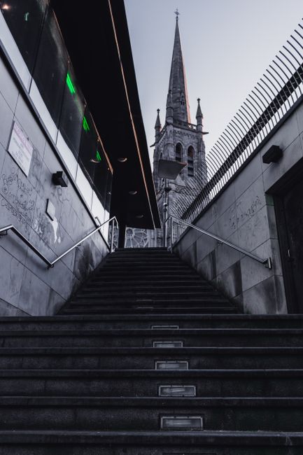 View from the bottom of an outdoor staircase leading up to a doorway, with metal handrails on both sides and stone steps in the foreground. To the left, part of a modern building façade with illuminated green indicator lights is visible. To the right, a black metal fence runs alongside the staircase. In the background, a historic church with a tall, pointed spire and Gothic architectural features, such as arched windows and decorative pinnacles, rises against a cloudy sky. The scene is lit with natural daylight, capturing the contrast between the contemporary urban environment and the historic church, illustrating a typical urban street scene that could be associated with home relocation or moving services like those offered by Man with Van St Mary Cray, especially during the process of transporting furniture or packing materials in a city setting.