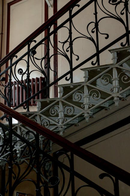 The image depicts a staircase inside a property, consisting of a white painted metal structure with decorative spiral and scroll patterns on the balustrade. The staircase has light grey wooden steps that lead upwards, with a polished dark wooden handrail running alongside. The stairs are situated beneath a high ceiling with a wooden-paneled wall in the background, featuring a section of a radiator partially visible at the top of the staircase. The environment appears to be a historic or Victorian-style building, with natural light illuminating the area. The scene is neutral, focusing on the architectural details of the staircase, which may be part of a home relocation or moving process managed by Man with Van St Mary Cray, as suggested by the context of the webpage related to removals.