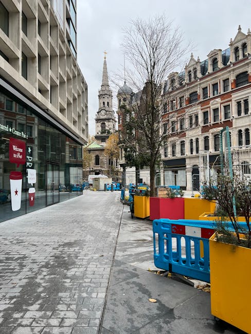 An urban roadside scene in Cray High Street, St Mary Cray, showing a paved pavement with several large, brightly coloured rectangular planters made of plastic or wood, painted in red, yellow, and blue, positioned along the edge of the sidewalk. Behind the planters, a leafless tree stands, surrounded by temporary blue safety barriers. To the left, the glass-fronted entrance of a modern building is visible, with signage and a red and white emergency call box attached to the exterior wall. In the background, historic church spires and domes rise above older, ornate residential buildings with bay windows, decorative facades, and wrought-iron balconies. The sky is overcast, and the street appears quiet with no pedestrians or vehicles presently visible. The scene is consistent with a street undergoing some traffic or pedestrian management related to house removals or moving logistics, with features typical of a local area in St Mary Cray. Man with Van St Mary Cray occasionally operates in this area for residential relocations, involving furniture transport, packing, and loading procedures.