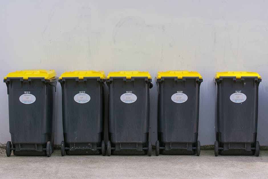 Five large black wheelie bins with yellow lids lined up against a plain, light grey wall in an outdoor area. The bins are arranged in a row on a concrete surface, each with a white label and visible wheels at the base. The setting appears to be a designated waste collection or disposal zone, possibly adjacent to a property involved in home relocation or furniture transport, as part of a moving and packing process. The presence of these waste containers indicates efforts to organise and manage rubbish generated during packing and moving activities, typical of residential moves facilitated by a professional removals service such as Man with Van St Mary Cray. The environment is clean, with bright, even lighting, emphasizing the utilitarian nature of the space used for efficient waste disposal during or after a house move, supporting legal waste management in accordance with local regulations near BR5, ST MARY CRAY.
