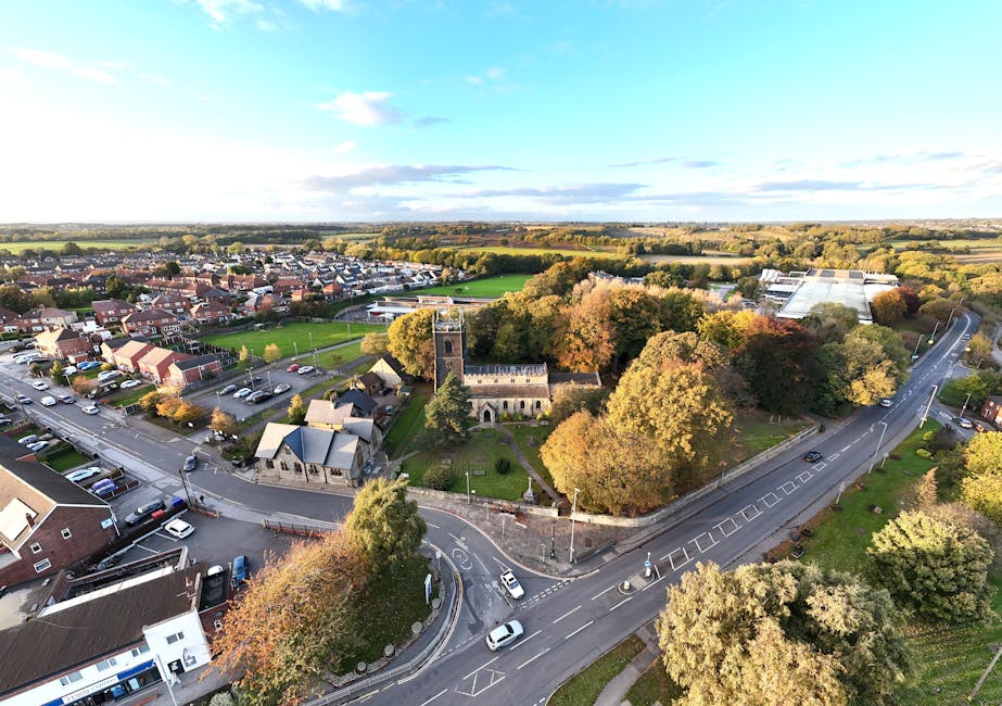 An aerial view of a small town area during daytime, featuring a church with a tall, narrow tower and a stone facade surrounded by a cemetery with green lawns and headstones. Adjacent to the church is a paved road with several cars parked along the curb and some vehicles in motion, including a small van near the church entrance. On the left side of the image, there is a parking lot filled with numerous parked cars next to residential houses with pitched roofs. To the right, the road curves around a collection of trees with autumn foliage, including shades of green, yellow, and orange, and continues into the distance over open fields with patches of farmland and countryside. The sky above is partly cloudy with patches of blue, and the lighting suggests late afternoon. This scene illustrates the process of house removals and furniture transport, with Man with Van St Mary Cray likely involved in the logistics of home relocation, moving furniture, and packing within this residential and community setting.